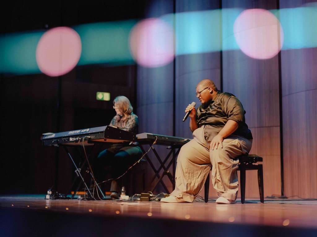 Zwei Musiker auf der Bühne: eine Person spielt Keyboard, die andere sitzt und singt in ein Mikrofon.