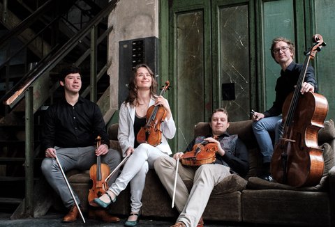Vier Musiker mit Streichinstrumenten sitzen und stehen in einem lässigen Ambiente. Sie wirken entspannt und bereit zu spielen.