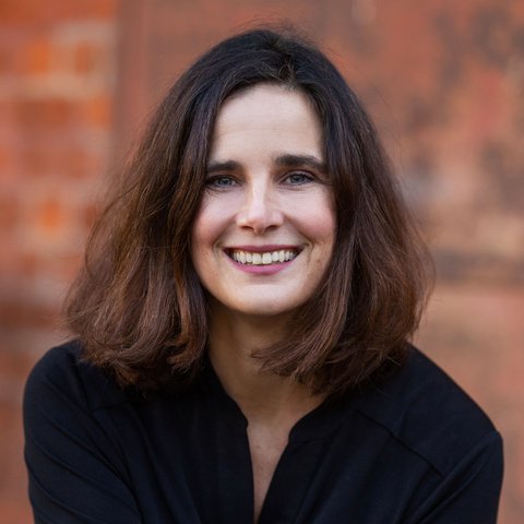 Eine Frau mit mittellangen braunen Haaren posiert in einem Porträtfoto für die Kamera. Sie trägt eine schwarze Bluse und lächelt.