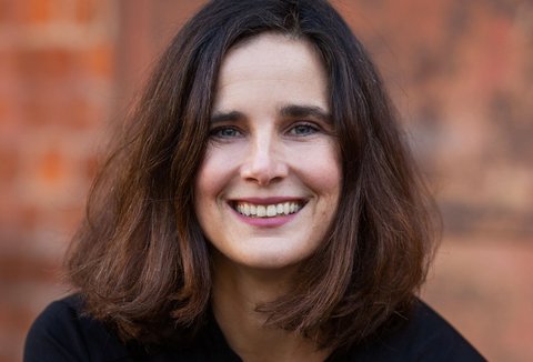 Eine Frau mit mittellangen braunen Haaren posiert in einem Porträtfoto für die Kamera. Sie trägt eine schwarze Bluse und lächelt.
