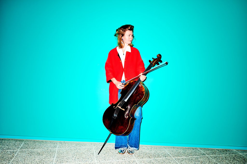 Eine Person in roter Jacke und Jeans hält ein Cello vor einem leuchtend türkisfarbenen Hintergrund. Kühler, minimalistischer Stil.