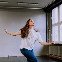 A woman dances passionately in a spacious room with large windows. She wears a white T-shirt and jeans.