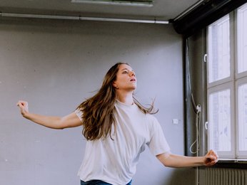 Eine Frau tanzt leidenschaftlich in einem geräumigen Raum mit großen Fenstern. Sie trägt ein weißes T-Shirt und Jeans.