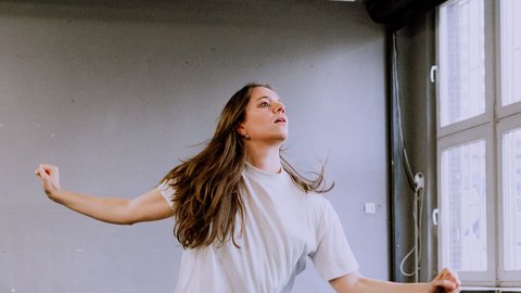 A woman dances passionately in a spacious room with large windows. She wears a white T-shirt and jeans.
