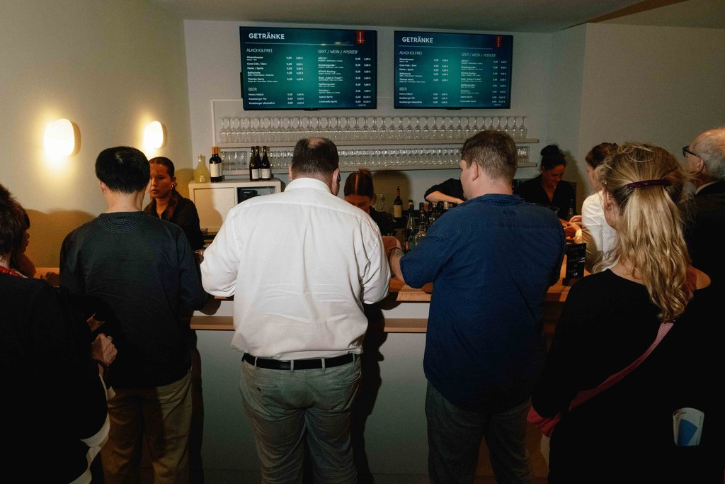 Menschen stehen an einer Theke in einer Bar oder einem Café. Menütafeln sind an der Wand hinter der Theke sichtbar.