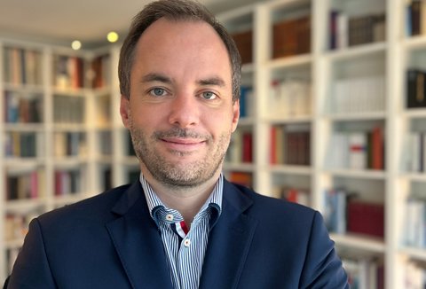 A man in a suit stands in front of a bookshelf. The shelves are filled with books, and the room is well-lit.