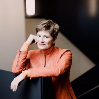 Eine Frau mit kurzen Haaren in einem roten Anzug posiert für die Kamera. Sie stützt den Kopf auf ihre Hand und lächelt.