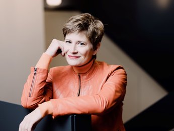 Eine Frau mit kurzen Haaren in einem roten Anzug posiert für die Kamera. Sie stützt den Kopf auf ihre Hand und lächelt.