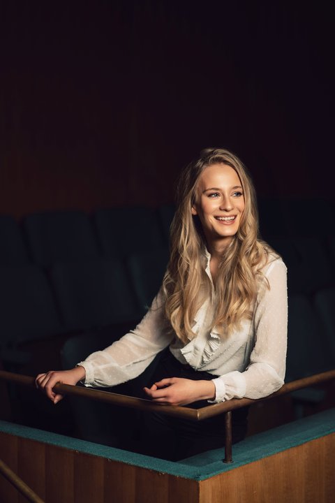 Eine Frau in einer weißen Bluse mit langen, blonden Haaren sitzt vor einem dunklen Hintergrund. Sie blickt nach rechts gedreht und lacht.