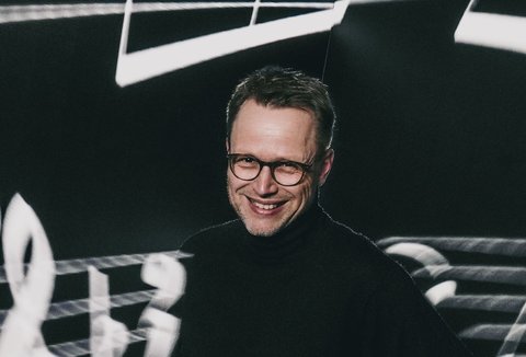 A man is standing in front of a dark background with stylised sheet music. He is wearing a dark sweater. He is looking into the camera and smiling. He has put one hand on his hip.