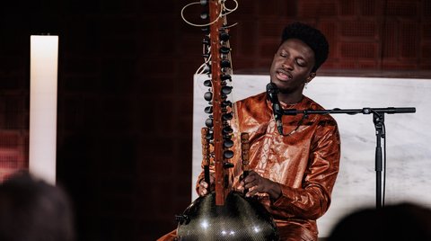Ein Musiker spielt konzentriert auf einer Kora, einem traditionellen westafrikanischen Saiteninstrument. Er trägt ein leuchtend oranges Outfit.