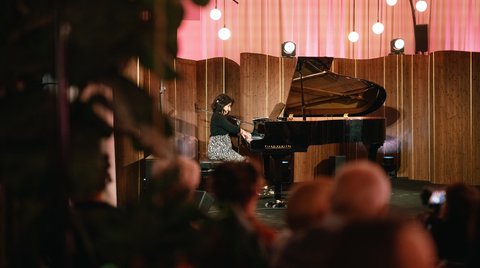 Frau spielt Klavier auf einer Bühne in einem stimmungsvoll beleuchteten Raum mit rosa Vorhängen. Zuschauer verfolgen das Konzert.