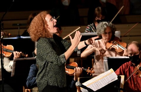Eine Dirigentin mit lockigem Haar leitet ein Orchester. Mehrere Musiker spielen Streicherinstrumente, konzentriert auf die Musik.