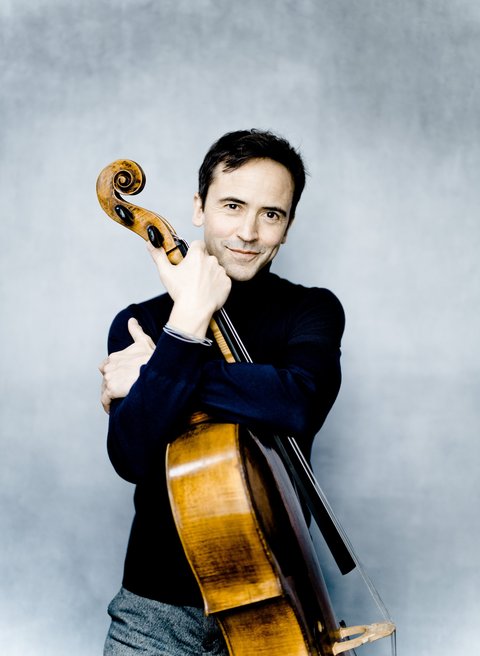 Ein Mann in dunklem Pullover umarmt ein Cello vor einem hellen, verschwommenen Hintergrund. Er lächelt sanft.