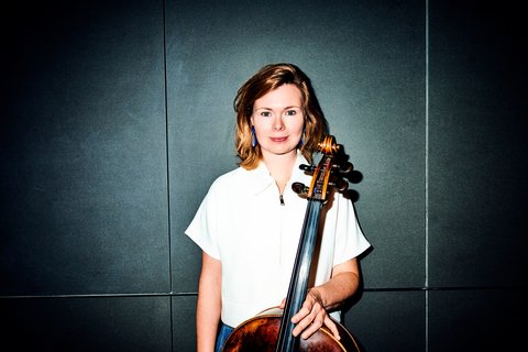 Frau in weißem Hemd hält ein Cello vor einem dunklen Hintergrund. Die Beleuchtung ist dramatisch und betont ihre Präsenz.