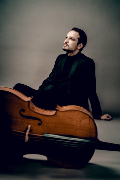 A man sits relaxed and contemplative, next to a large cello, in a professional studio setting.  © Andrej Grilc
