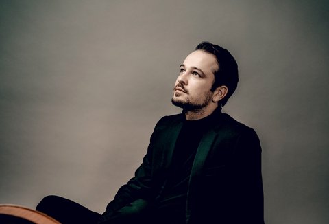 A man sits relaxed and contemplative, next to a large cello, in a professional studio setting.