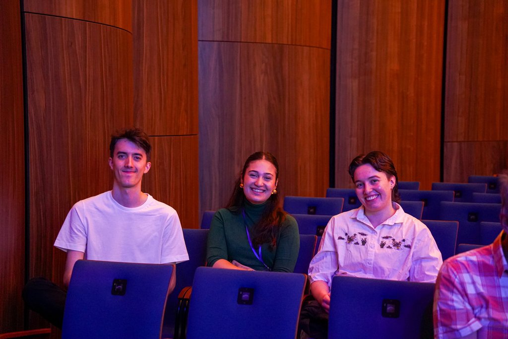 Drei Personen sitzen in einem Auditorium auf blauen Stühlen und lächeln. Sie scheinen eine Veranstaltung zu genießen.