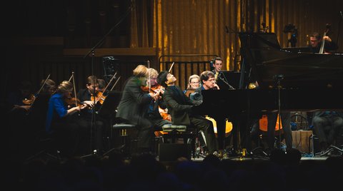 Ein Orchester spielt auf einer Bühne in einem Konzertsaal. Musiker konzentrieren sich auf ihre Instrumente, mit einem Flügel im Vordergrund.