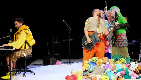 Three performers in colorful costumes on stage, playing music, surrounded by a pile of plush toys. © Claus Overkamp