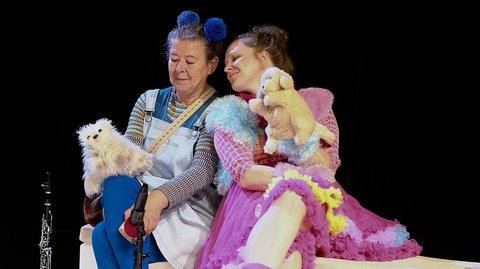 Two actors in colorful costumes, one in blue and the other in pink, holding plush toys while sitting on stage. © Claus Overkamp