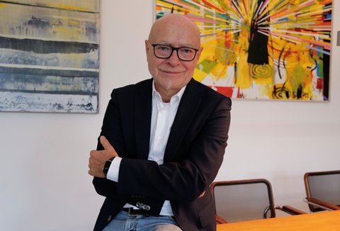 A man is standing in front of several pieces of art. He is wearing a suit. His arms are crossed. He is looking into the camera and smiling.