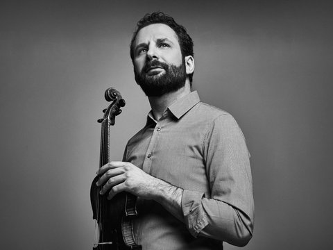 Ein Mann posiert mit einer Violine. Er trägt ein Hemd und blickt in die Ferne.