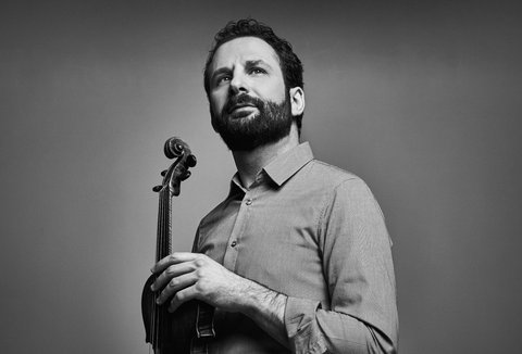 Ein Mann posiert mit einer Violine. Er trägt ein Hemd und blickt in die Ferne.