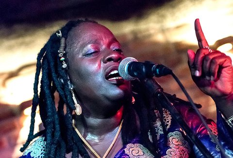 Eine Frau mit langen Dreadlocks singt leidenschaftlich in ein Mikrofon auf der Bühne. Der Hintergrund ist stimmungsvoll beleuchtet.