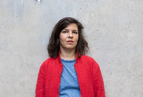 A woman is posing in front of a light wall. She is wearing a t-shirt and a jacket and looking into the camera.