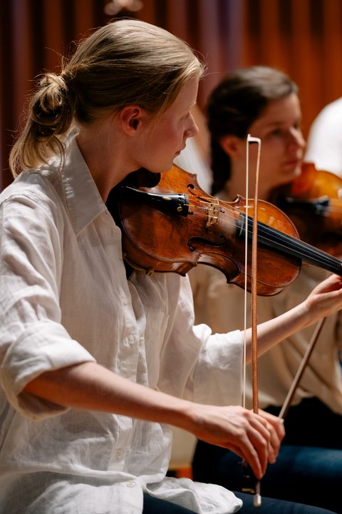 Zwei Geigerinnen spielen konzentriert bei einem Konzert. Sie tragen weiße Blusen und sind tief in ihre Musik vertieft.