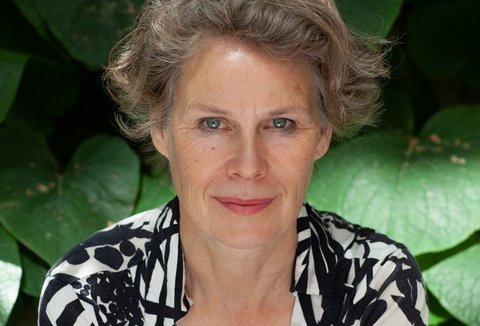 Eine Frau posiert vor den Blättern einer Pflanze. Sie trägt eine gemusterte Bluse, blickt in die Kamera und lächelt.
