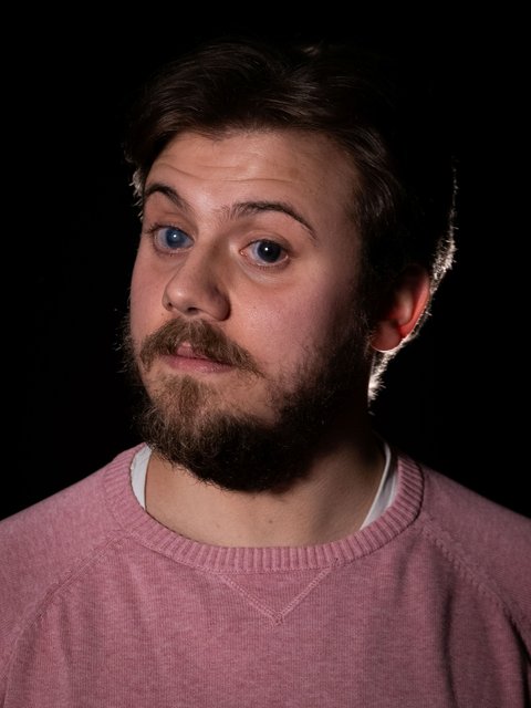 Portrait von einem bärtigen Mann in einem roten Pullover. Er posiert vor einem schwarzen Hintergrund und blickt in die Kamera.