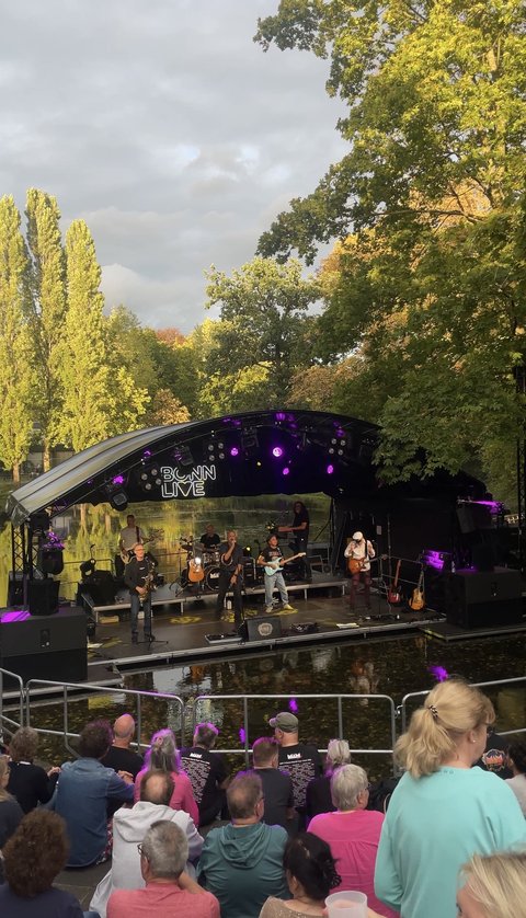 Ein Freiluftkonzert in einem Park mit einer Band auf einer Bühne. Zuschauer sitzen auf Bänken. Bäume im Hintergrund. ©RheinEvents Konzerte GmbH