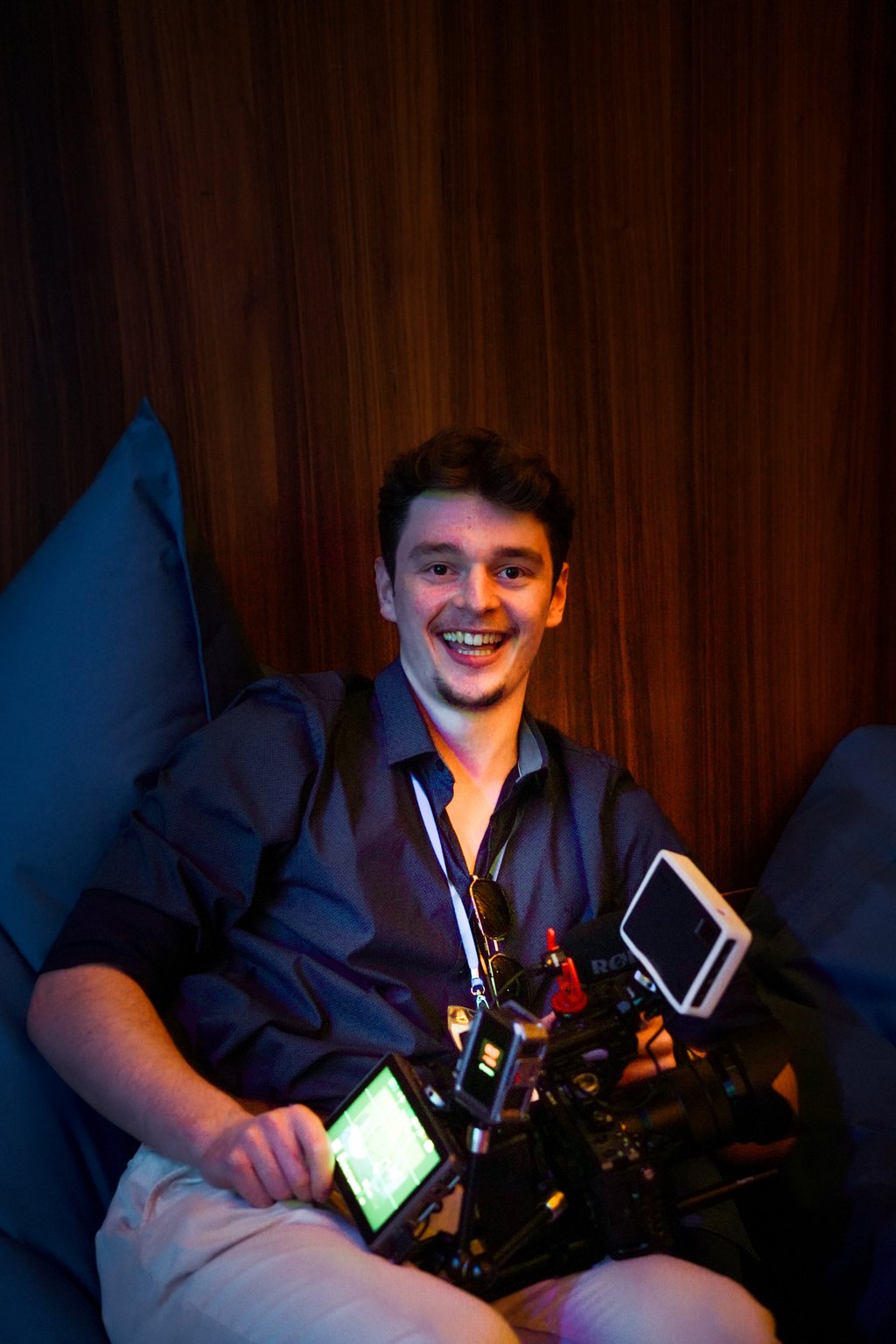 A man sits on a chair holding a film camera setup and smiling. The background features a wooden wall and cushions.