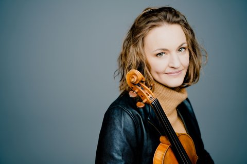 A woman with a violin, wearing a leather jacket. The background is light gray. She has a gentle smile and medium-length hair.