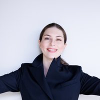A woman in a dark suit smiles with arms outstretched. She stands against a light background.