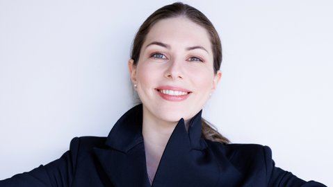 A woman in a dark suit smiles with arms outstretched. She stands against a light background.