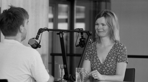Eine Frau und ein Mann sitzen sich an einem Tisch gegenüber. Zwischen ihnen steht Podcast-Equipment.