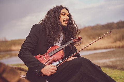 A person with long hair in a coat sits outdoors with a violin. The background features a natural landscape.