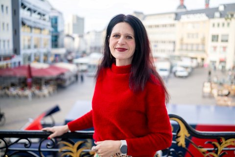 Eine Frau in einem roten Pullover posiert vor dem Bonner Markt. Sie lächelt und blickt in die Kamera.