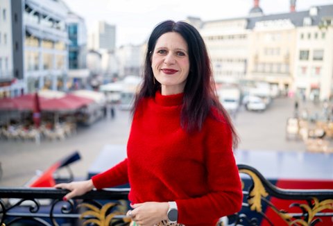 Eine Frau in einem roten Pullover posiert vor dem Bonner Markt. Sie lächelt und blickt in die Kamera.