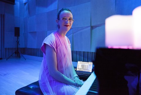 A person in pink clothing playing piano in a blue, mood-lit room with candles in the foreground.
