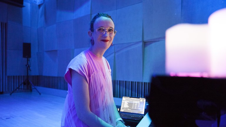 A person in pink clothing playing piano in a blue, mood-lit room with candles in the foreground.