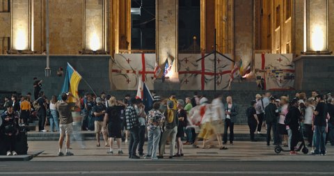 Eine Gruppe von Menschen versammelt sich vor einem großen, beleuchteten Gebäude. Einige halten Flaggen, darunter eine in Blau und Gelb.