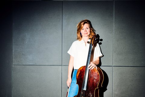 Eine Frau steht mit einem Cello in einem Raum mit grauen Wänden. Sie trägt ein weißes Hemd und Jeans.