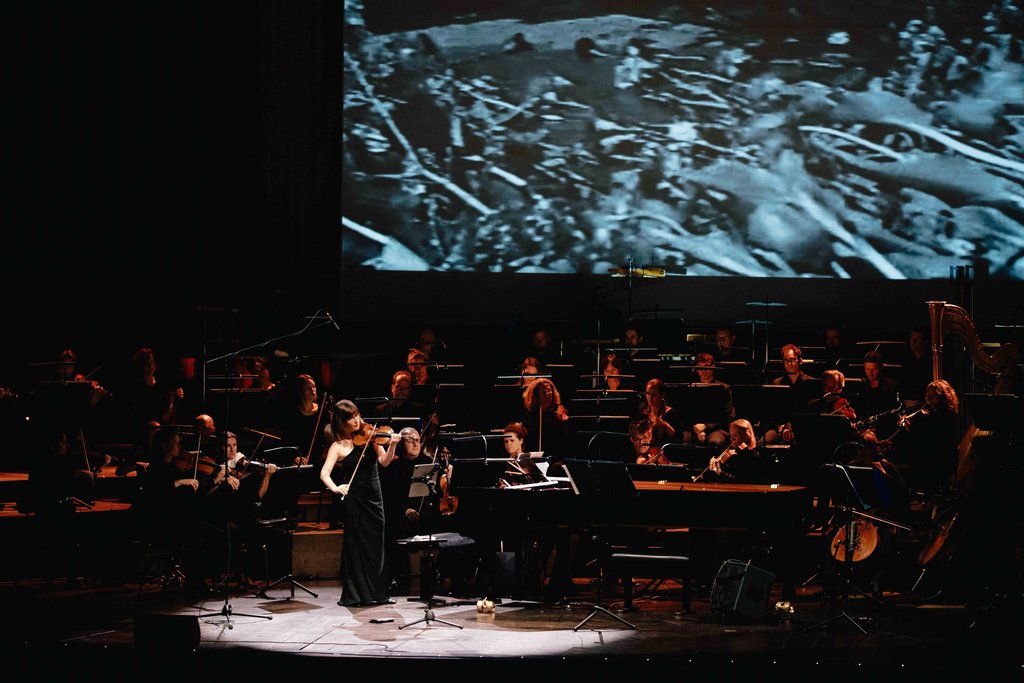 Eine Musikerin spielt Flöte auf einer Bühne vor einem Orchester, während ein schwarz-weißes Bild auf die Leinwand projiziert wird.