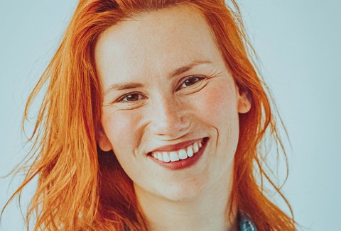 A woman is standing in front of a light background. She is wearing a patterend shirt. She is looking into the camera and smiling.