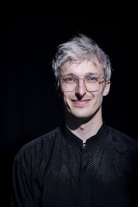 Ein Mann mit hellem Haar und Brille in dunkler Kleidung lächelt in einem dunklen, minimalistischen Hintergrund.