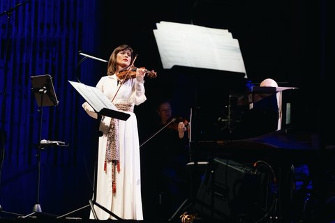 Eine Violinistin in einem weißen Kleid spielt leidenschaftlich auf der Bühne. Notenständer und Orchester im Hintergrund erkennbar.